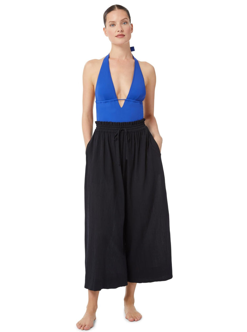 Model posing with her hands in her pockets wearing a black organic cotton beach pants with elastic waistband with drawstring and tassel detail with a cobalt blue deep plunge one piece bathing suit