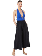 Model posing with her hands in her pockets wearing a black organic cotton beach pants with elastic waistband with drawstring and tassel detail with a cobalt blue deep plunge one piece bathing suit