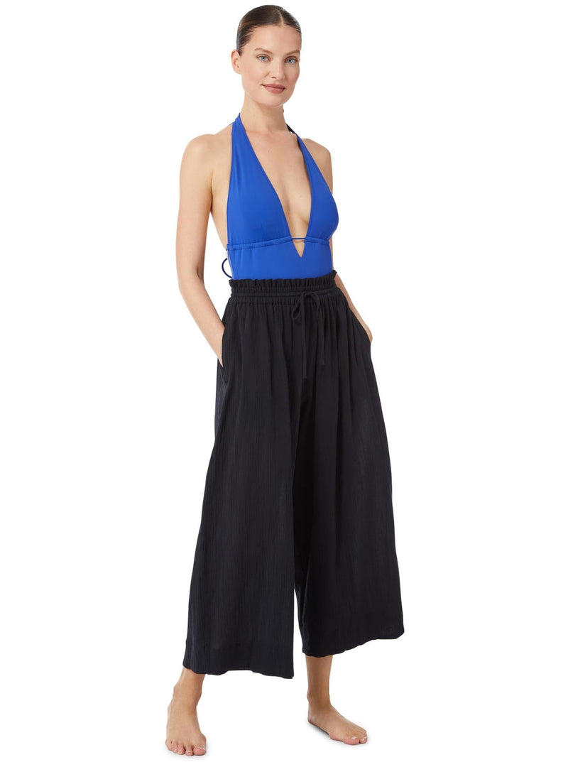 Model posing with her hands in her pockets wearing a black organic cotton beach pants with elastic waistband with drawstring and tassel detail with a cobalt blue deep plunge one piece bathing suit