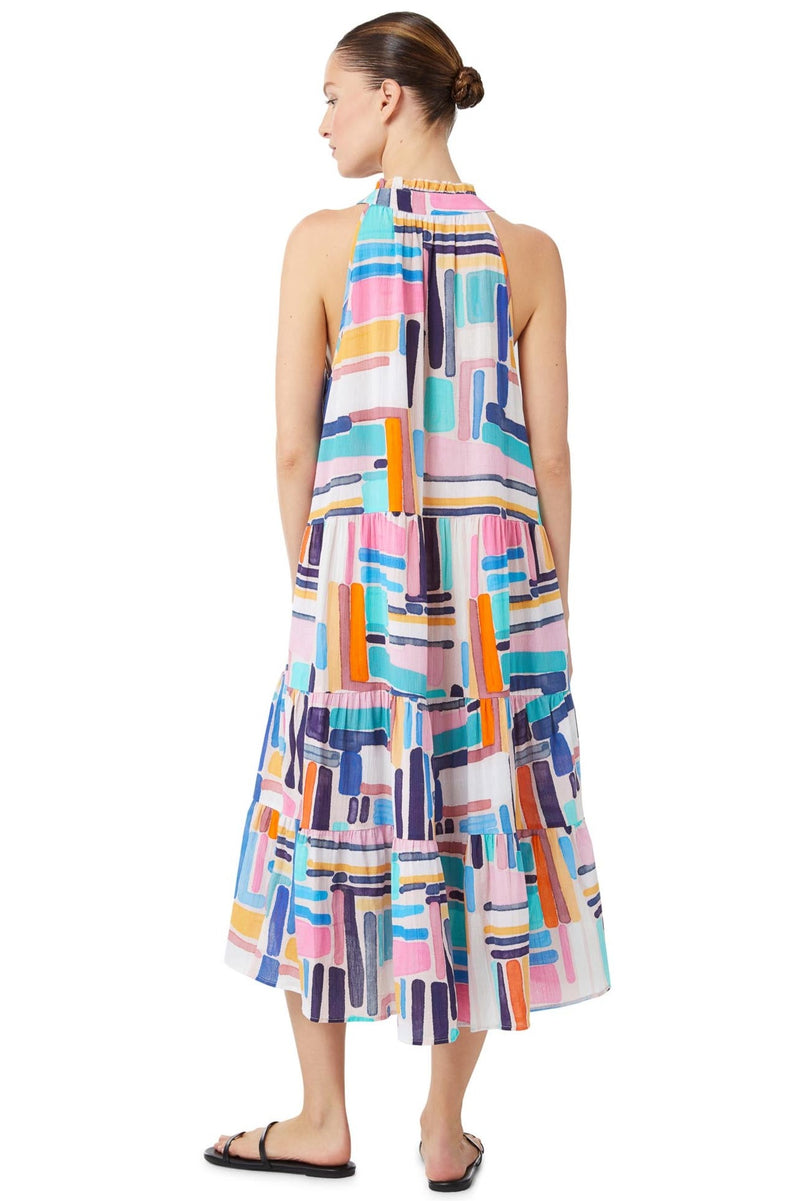 The back of a model wearing a geometric and graphic colored sleeveless, high neck with ruffle detail, buttoned shirt dress with optional matching belt with tassels with hands in pockets. 