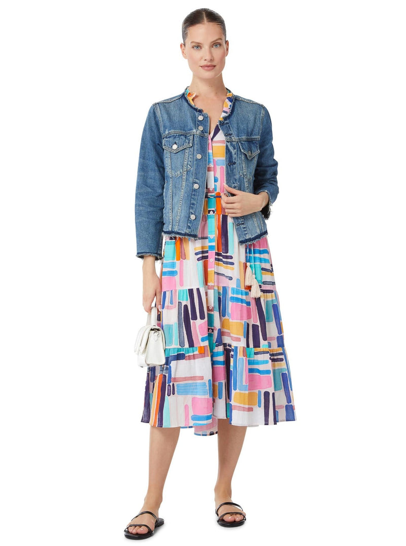 Model wearing a geometric and graphic colored sleeveless, high neck with ruffle detail, buttoned shirt dress with optional matching belt with tassels and pockets, styled with a denim jacket, white purse, and black sandals. 