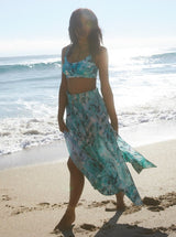Model walking on beach wearing ocean print inspired midi length skirt with side slits and elastic waistband with drawstring detail with on side seam pockets with matching ocean print bikini top