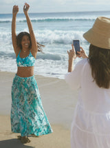 Model in fresh white dress and straw sun hat talking a photo of a model on the beach posing with her hands up wearing ocean print inspired midi length skirt with side slits and elastic waistband with drawstring detail with pockets and matching ocean print bikini top