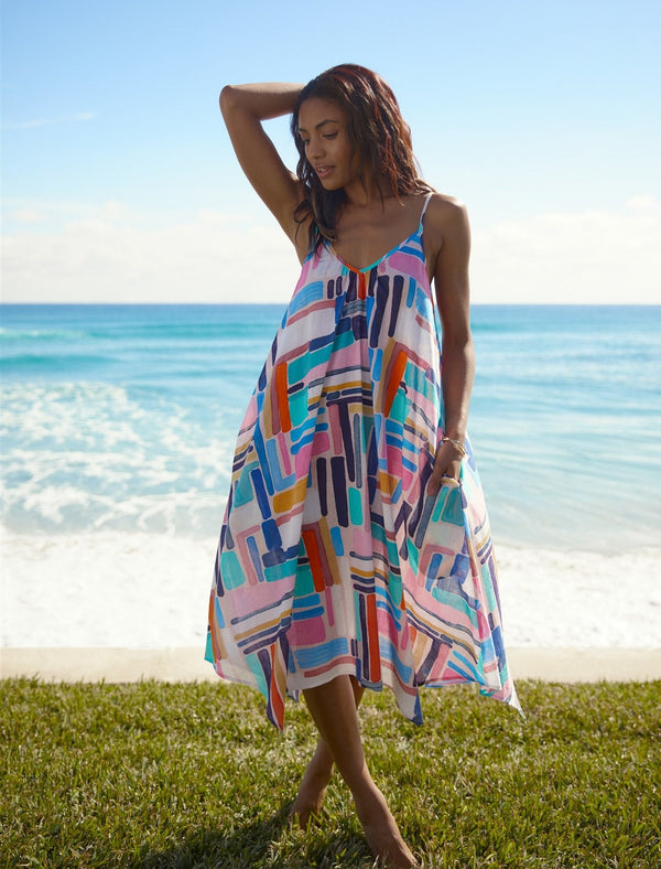 Model standing in front of a beach with hands behind her head wearing a geometric and graphic colored strappy and long flowy dress with adjustable back shoulder straps, v-neckline front and pockets 
