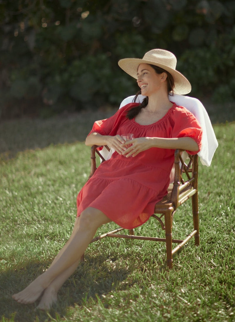 model laughing and sitting on a chair in the lawn wearing a coral red slightly above the knee dress with rounded neckline and short puff sleeve with a covered elastic cuff and a straw hat