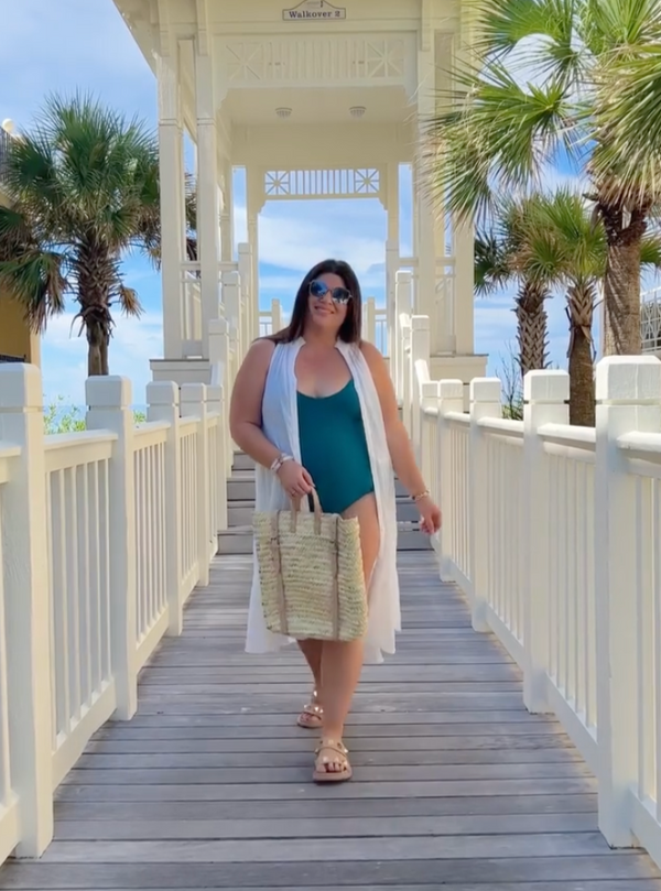 Women standing on beach dock in front of palm trees wearing Green v-neckline one piece bathing suit with adjustable back straps with white duster dress, straw beach bag, and sunglasses