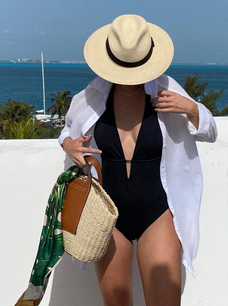 model wearing black deep plunged one piece swimsuit with an adjustable halter-neck tie and under-bust string with linen button up shirt, straw hat, and straw purse