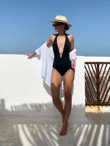 model wearing black deep plunged one piece swimsuit with an adjustable halter-neck tie and under-bust string with linen button up shirt, straw hat, and sunglasses